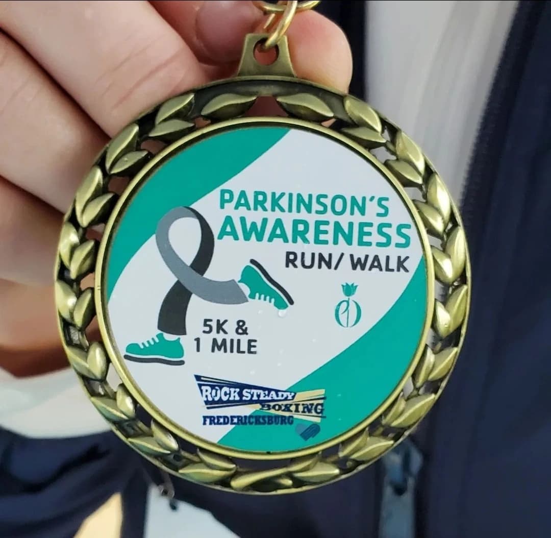 A hand holds a Parkinson’s Awareness Run/Walk medal bearing the Rock Steady Boxing Fredericksburg logo.