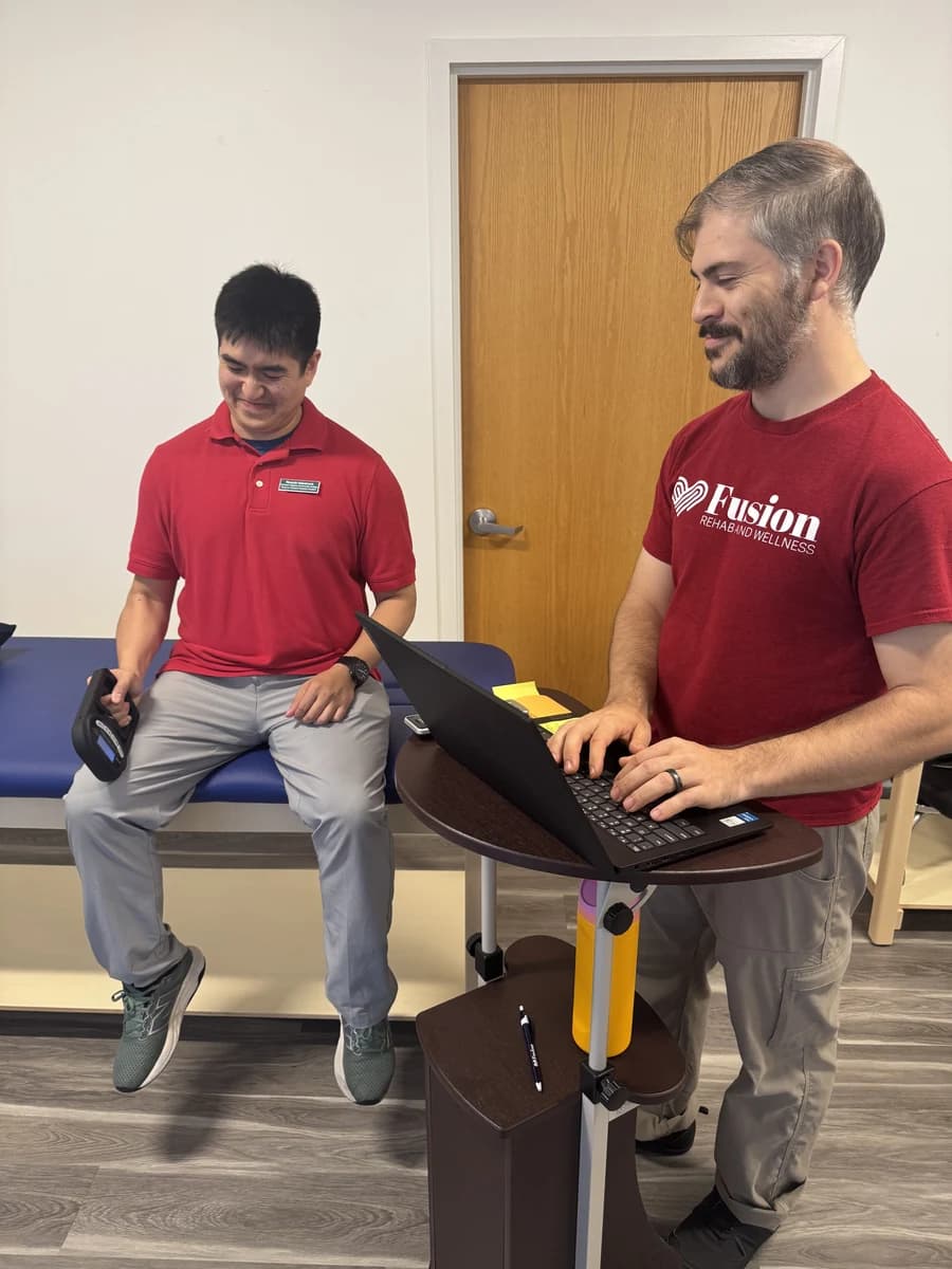 A seated man holds a grip-testing device while a Fusion therapist records information.