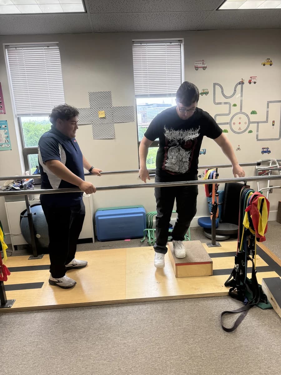 A man practices stepping onto a low platform between parallel bars with a Fusion therapist beside him.